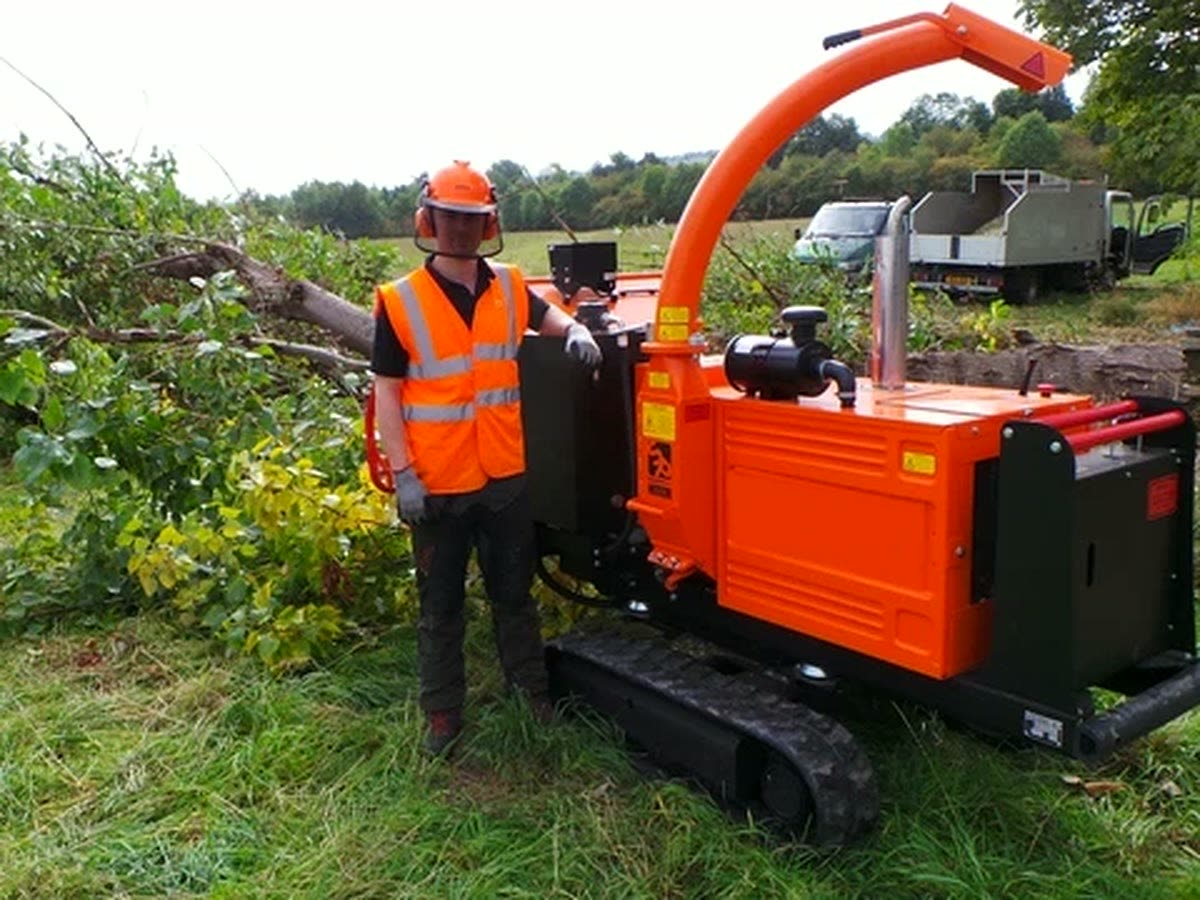Operator in hi-vis and helmet beside a tracked wood chipper with a pile of branches